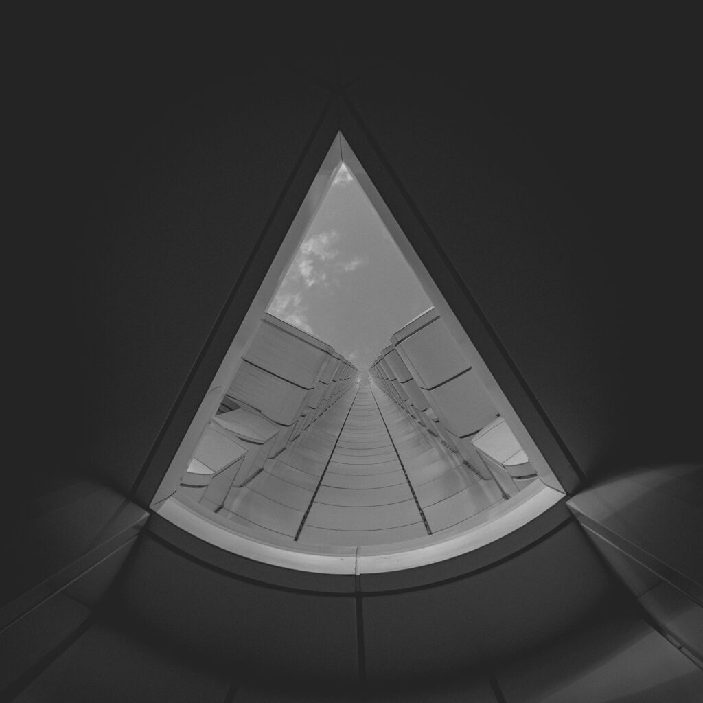 Himmelsspalt Durch ein architektonisches Dreieck öffnet sich der Blick senkrecht nach oben – zwischen klaren Linien und rhythmischer Fassadenstruktur zieht die Perspektive den Blick in die Tiefe des Himmels. Der Moment zwischen Geometrie und Unendlichkeit, in kontrastreichem Schwarzweiß gehalten, betont die skulpturale Wirkung des Bauwerks.