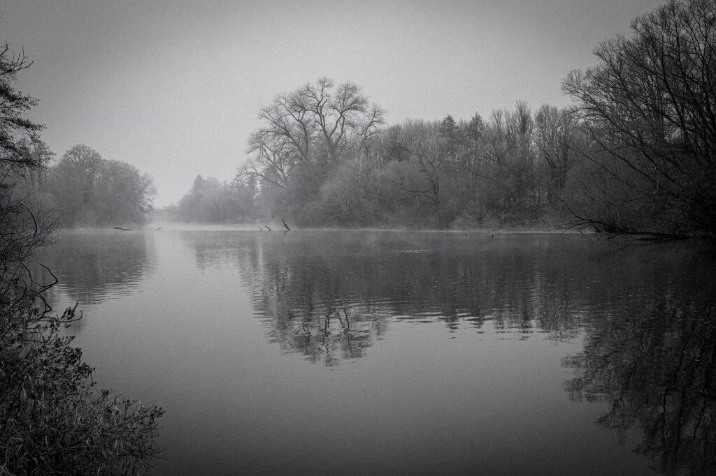 Reflextion Stille Momentaufnahme an der Lahn: Im Nebel liegt das Wehr fast verborgen, die Wasseroberfläche spiegelt die winterlich kahlen Bäume in sanften Wellen. Eine Szene voller Ruhe und Tiefe, festgehalten in Schwarz-Weiß. Das Bild lädt ein, innezuhalten – ein Blick in die Landschaft wird zum Blick nach innen.