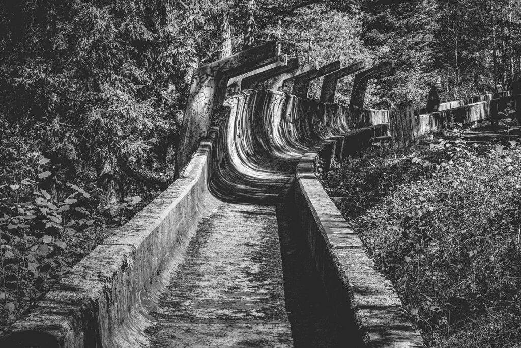Vergessene Spuren Eingebettet in die dichten Wälder des Trebević-Gebirges schlängelt sich die einstige Bobbahn von Sarajevo durch die Landschaft – ein Relikt der Olympischen Winterspiele von 1984. Einst ein Ort des sportlichen Wettkampfs, ist sie heute von der Natur zurückerobert und von der Zeit gezeichnet. Die verwitterten Betonwände, überzogen von Graffiti und Moos, erzählen von vergangenen Zeiten und neuen Geschichten. Dieses Fine Art Print fängt die raue Schönheit und die Melancholie dieses Ortes in einer eindringlichen Schwarz-Weiß-Komposition ein – ein stilles Echo der Geschichte.