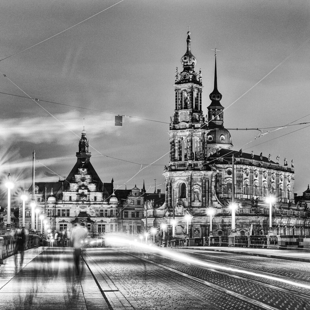 Schwarzweißaufnahme der Dresdner Hofkirche bei Abendstimmung, aufgenommen von der Augustusbrücke. Langzeitbelichtung mit Lichtspuren vorbeifahrender Fahrzeuge und verschwommenen Fußgängern im Vordergrund.