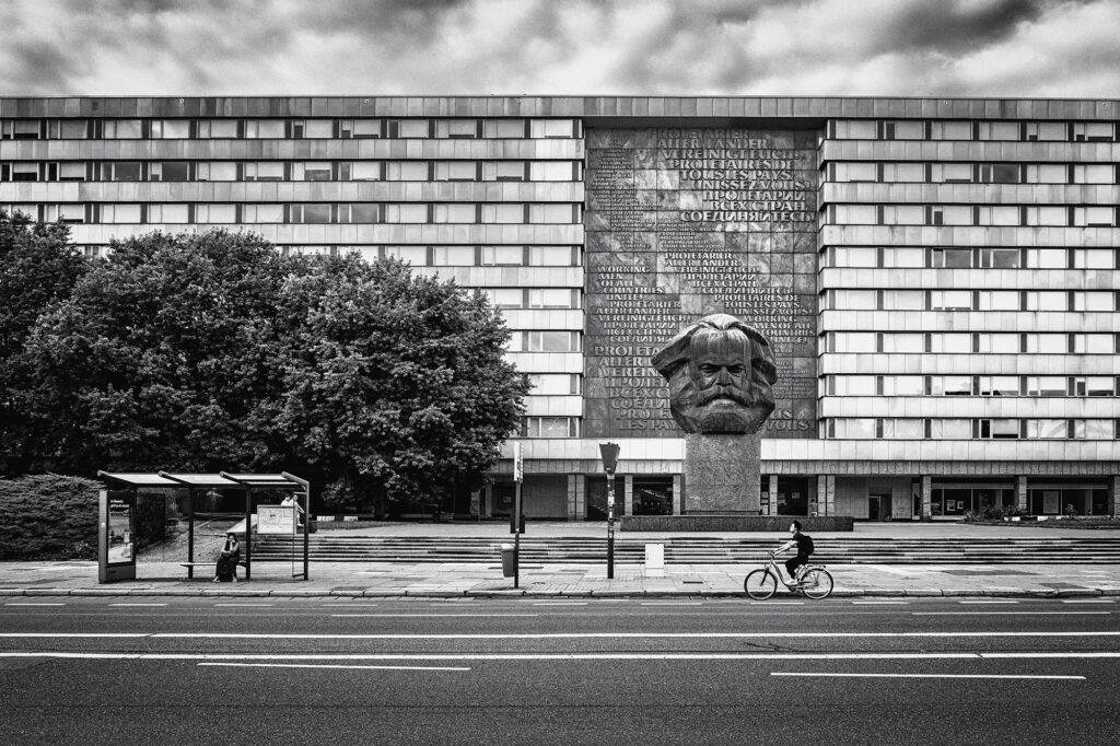 Unser Bestreben war es, die urbane Gelassenheit dieser Szene festzuhalten, indem wir den hektischen Straßenverkehr ausblenden. Wir haben die Menschen beobachtet, die kommen und gehen, und diesen einen perfekten Moment eingefangen. Inmitten der Hektik des städtischen Lebens entstand ein Moment der Ruhe und Gelassenheit vor der beeindruckenden Kulisse der Karl Marx Statue in Chemnitz. Diese Streetart-Szene erinnert uns daran, wie faszinierend und unvorhersehbar das urbane Leben sein kann.