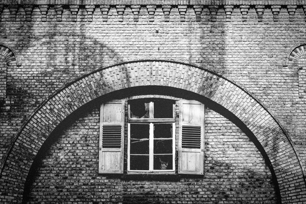 Fensterblick Die Schwarz-Weiß-Aufnahme fängt die Textur und Patina der Horchheimer Torbefestigung eindrucksvoll ein. Ein einzelnes Fenster mit gebrochenen Scheiben und verwitterten Fensterläden steht im Zentrum, umrahmt von den klaren Linien des steinernen Bogens. Die Bildkomposition betont den Kontrast zwischen der beständigen Architektur und der vergänglichen Zeit, die sich in den Spuren am Fenster zeigt. Die monochrome Darstellung unterstreicht die zeitlose Eleganz und die beeindruckende Ausdauer des Bauwerks.