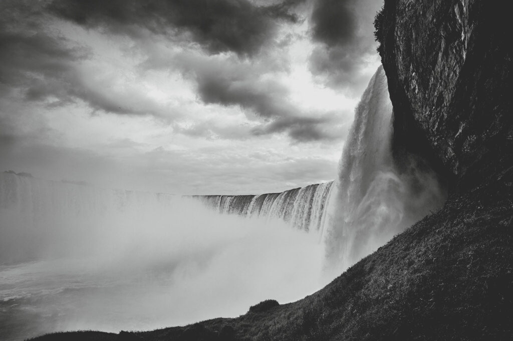 niagara – behind the falls Das Kunstwerk "Niagarafälle - Behind the Falls" zeigt eine außergewöhnliche Sicht auf die eindrucksvollen Niagarafälle – von unten betrachtet. In Schwarz-Weiß-Fotografie eingefangen, wird die mächtige Schönheit des Naturwunders verdeutlicht, wenn die Wassermassen majestätisch über die Felswand hinabstürzen. Diese gewaltige Darstellung kommt auf einem großen GalleryPrint besonders eindrucksvoll zur Geltung.