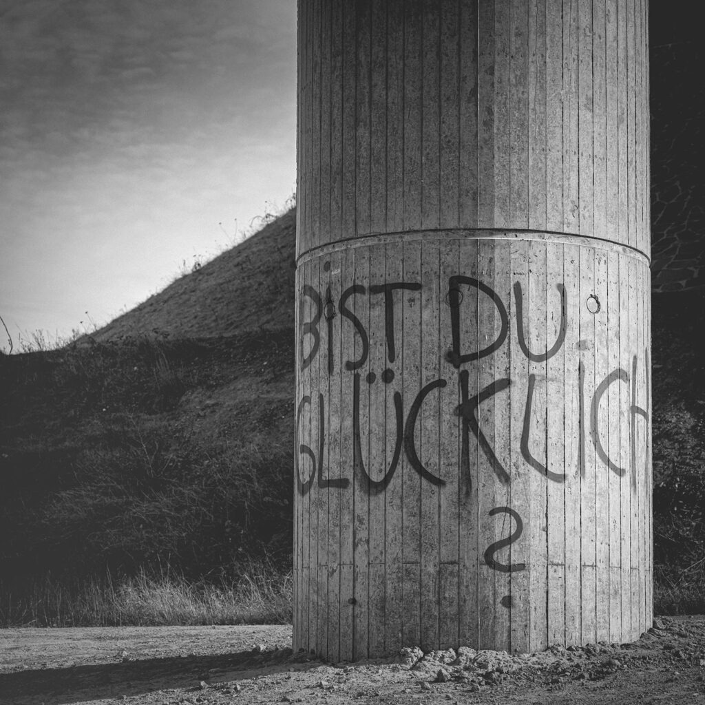 Bist du Glücklich Schwarzweißfotografie einer Betonstütze mit Graffiti-Aufschrift „Bist du glücklich?“, die zum Nachdenken über das eigene Leben anregt