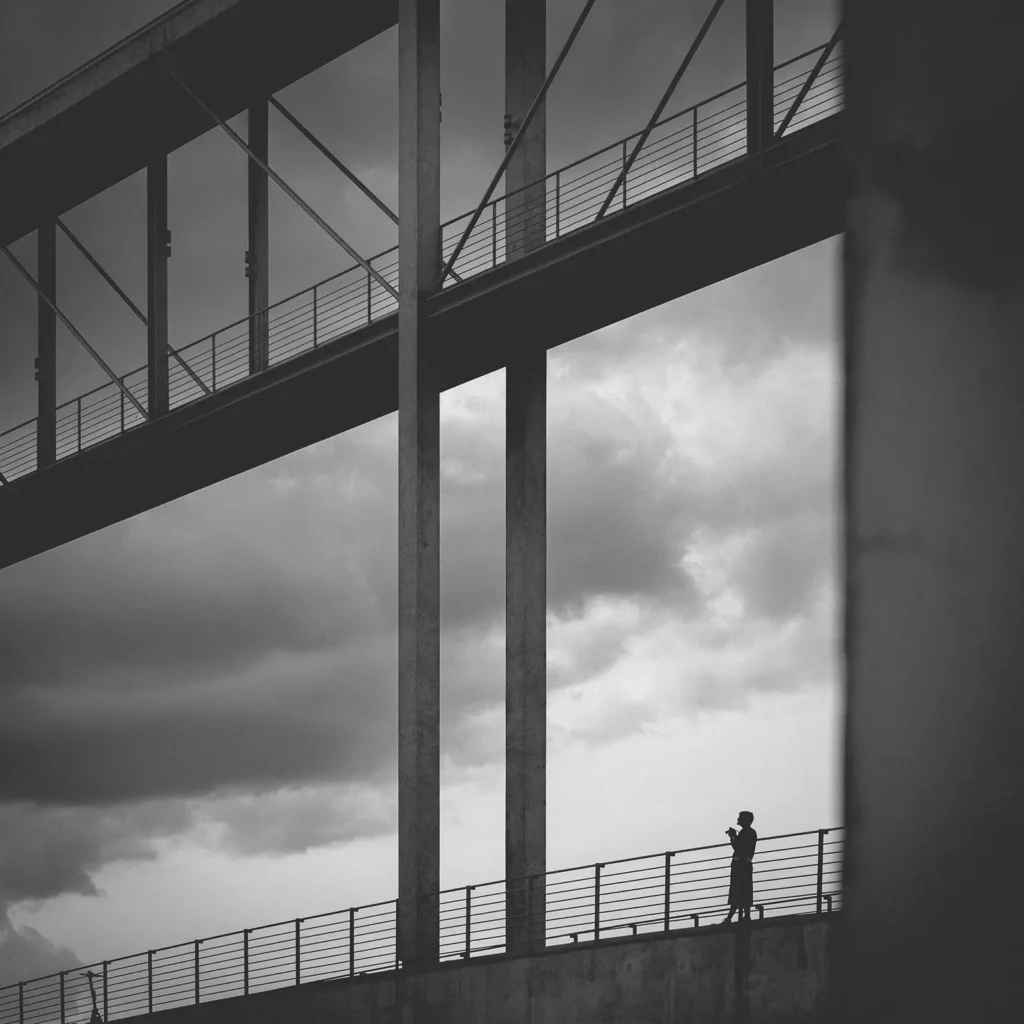 Schwarzweißfotografie einer modernen Fußgängerbrücke mit klaren Linien und geometrischen Strukturen, ein Mensch als kleine Silhouetten.