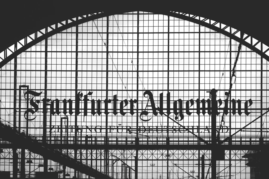 Schwarzweißaufnahme des Frankfurter Hauptbahnhofs mit dem Schriftzug „Frankfurter Allgemeine“ auf dem Glasdach.