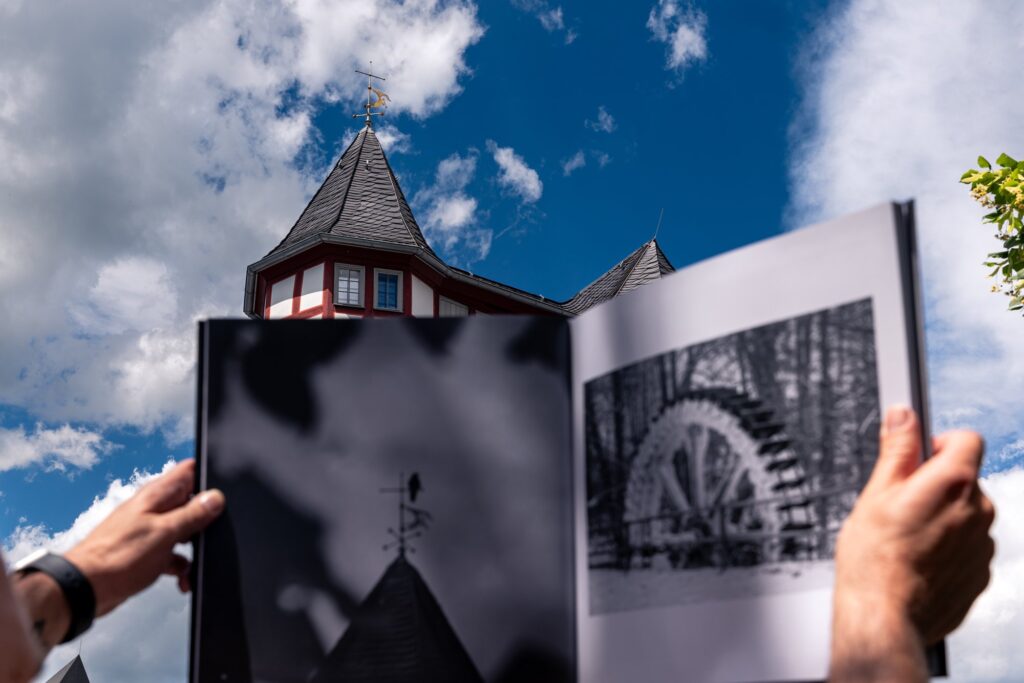 Hände halten den Bildband „Limburg – Zeitlos in Schwarz & Weiß“ mit Wetterfahne und Mühlrad vor blauem Himmel